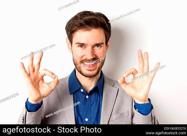 Smiling businessman in suit showing okay signs in approval, guarantee quality product, praise good thing, standing on white background