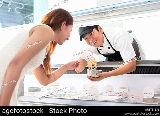 Helpful young sales assistant smiling at a customer through the glass of the display counter as he leans forwards to serve her selection of food
