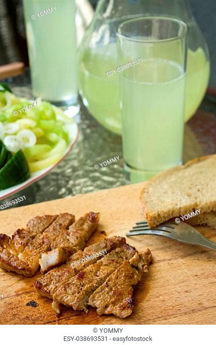 Vertical photo of pork steak cut on stripes placed on chopping board with fork and bread. Vegetable and cucumber lemonade in glass and jar are around