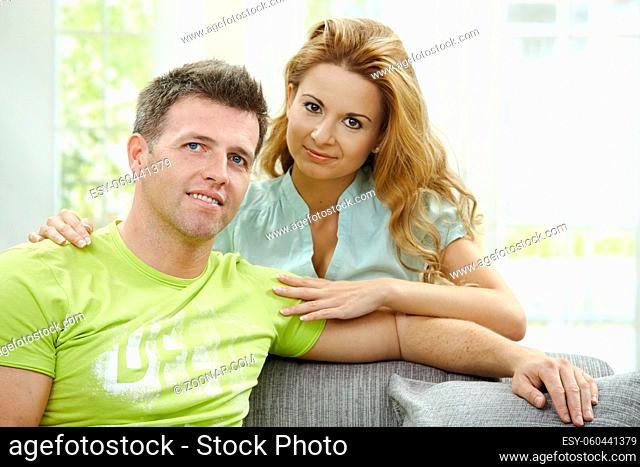 Love couple at home. Man sitting on sofa, his girlfriend embracing him from behind