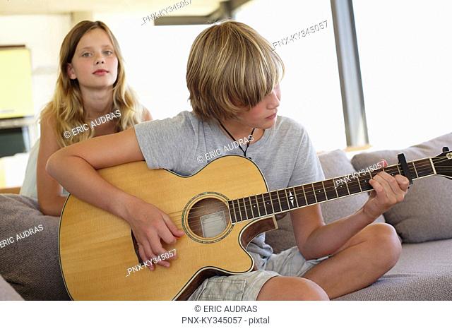 Teenage boy playing a guitar with her sister standing behind him listening