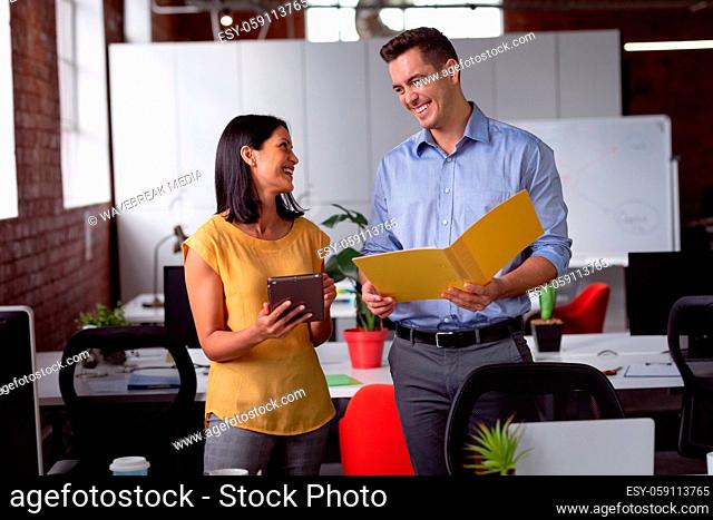 Happy caucasian male and female colleague standing at desk holding tablet and folder and smiling
