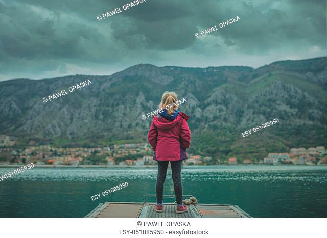 Caucasian girl in a waterproof red jacket standing on the pier in Kotor Bay, Montenegro