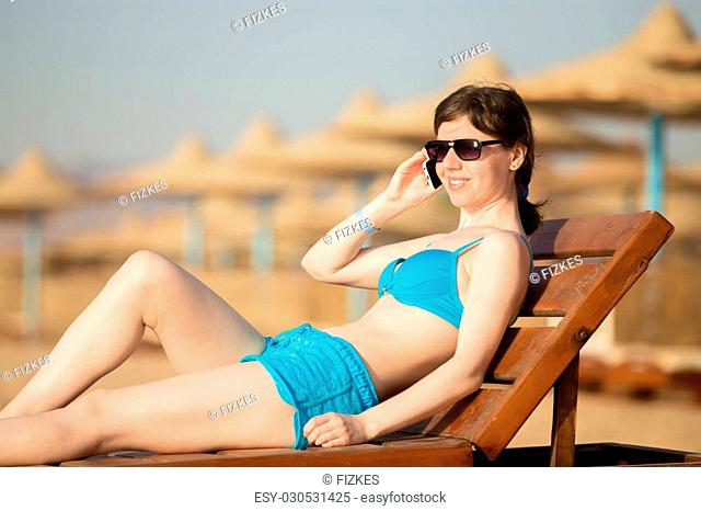 Young slim happy smiling woman in sunglasses relaxing and tanning on wooden sun lounger on sunny beach with straw umbrellas, making call on cell phone