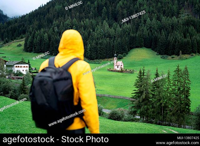 Hiking man with backpack enjoy the view of St. Johann church in Val di Funes valley, Dolomites, Italy, Europe