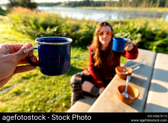 Selective focus of two people cheering their cups of coffee or tea while having a picnic on a sunny day outdoors in a nature scenery