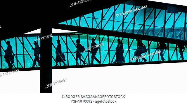 Passengers disembark from an aeroplane through the enclosed passage ramp. They are silhouetted against the sunlight
