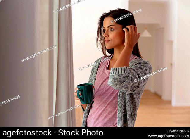 Caucasian woman standing by window at home drinking cup of coffee and smiling