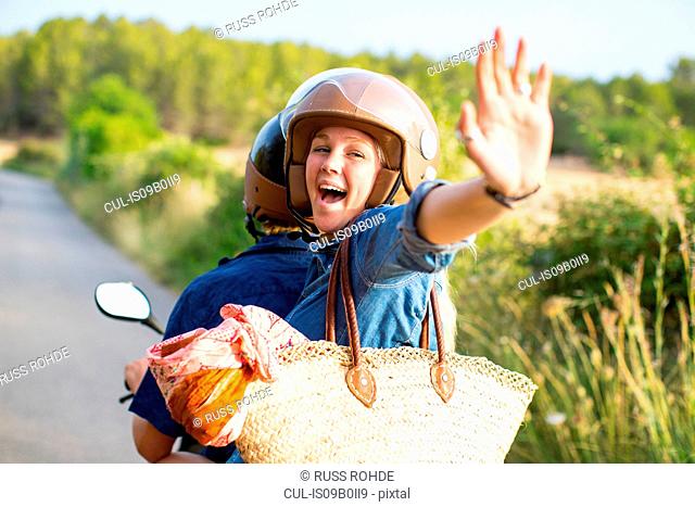 Young woman riding pillion on rural road waving, Majorca, Spain