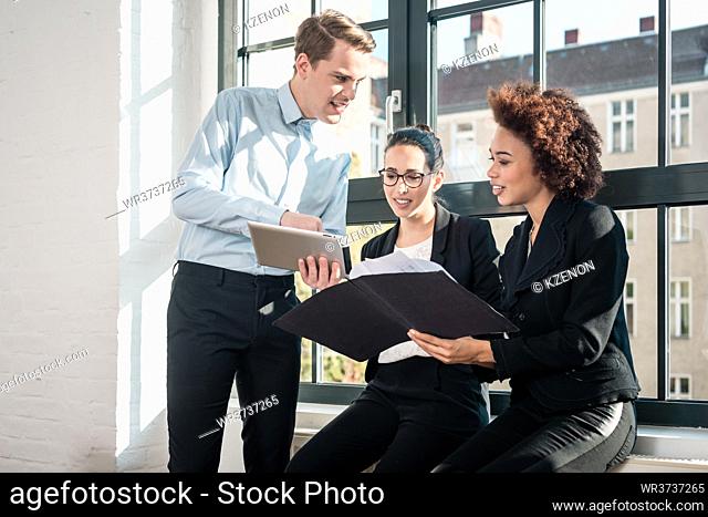 Three young cheerful employees smiling while holding a tablet PC and a folder during break in a modern office