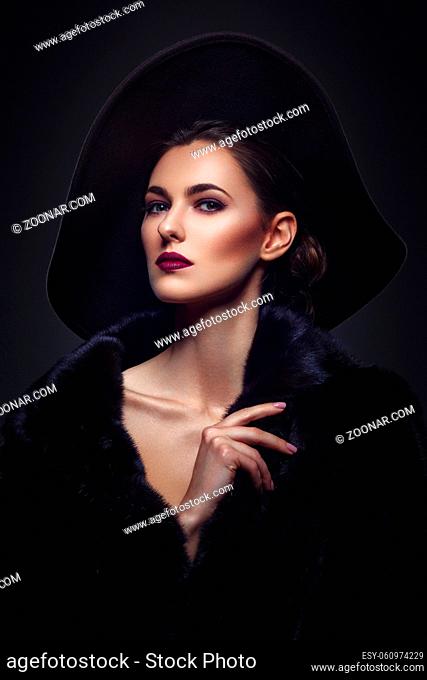 Beautiful young woman with glamorous make-up in luxurious fur coat and fashion hat on black background. Copy space