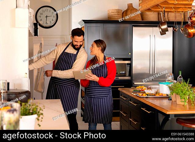 Happy caucasian couple preparing food, smiling and looking at recipe on tablet in kitchen
