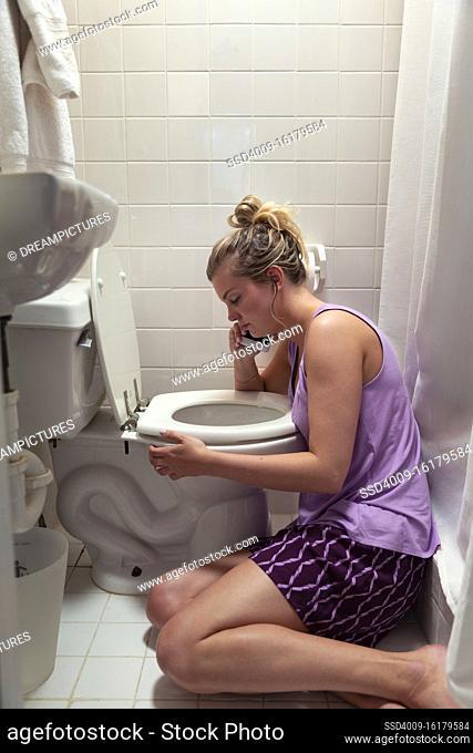 Young woman on the floor in her bathroom feeling sick next to toilet, talking to doctor on cellphone