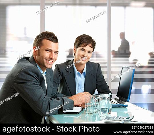 Businessman and businesswoman sitting at desk in office, using laptop computer, smiling