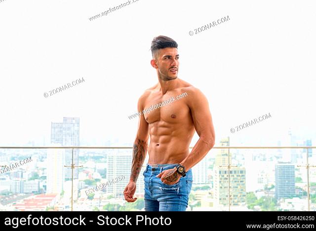 Portrait of young handsome muscular Persian man shirtless and looking macho against view of the city