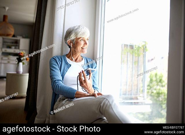 Active senior woman listening to music on smart phone while looking out of window sitting at home