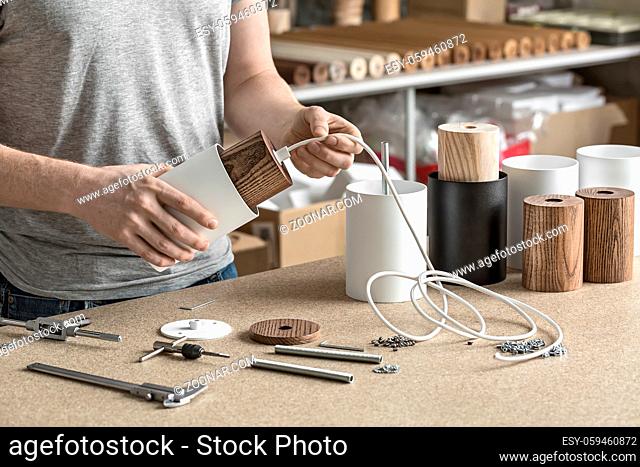 Lamp assembling in the workshop. Man holds a white lamp with a wooden part per the cable. On the table there are other lamp billets, different wrenches, caliber