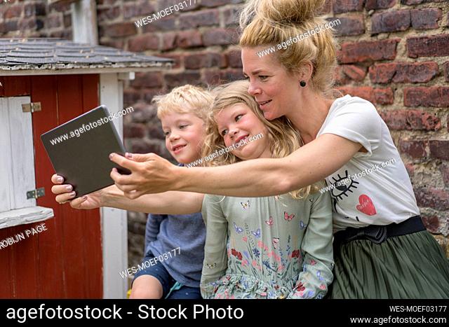 Woman taking selfie with kids through digital tablet against brick wall at back yard