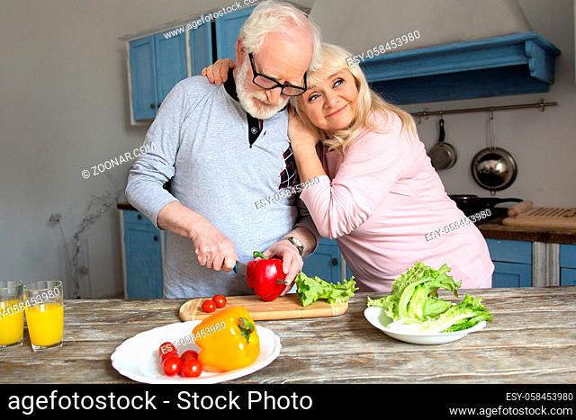 Charming aged man and woman enjoy spending time together in kitchen. Elderly man cutting red bell pepper for lunch with his wife hugging him with modern...