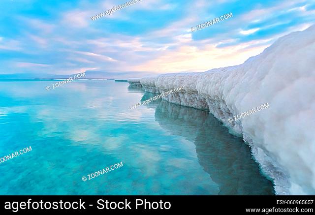 Sand completely covered with crystalline salt looks like ice or snow on shore of Dead Sea, turquoise blue water near, sky colored with morning sun distance -...