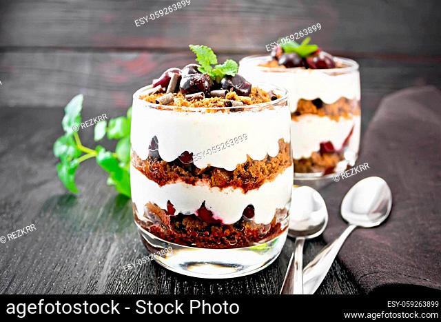 Dessert Black Forest of cherries, chocolate biscuit and soft cottage cheese with cream in two glasses, a napkin, mint and spoons on wooden board background