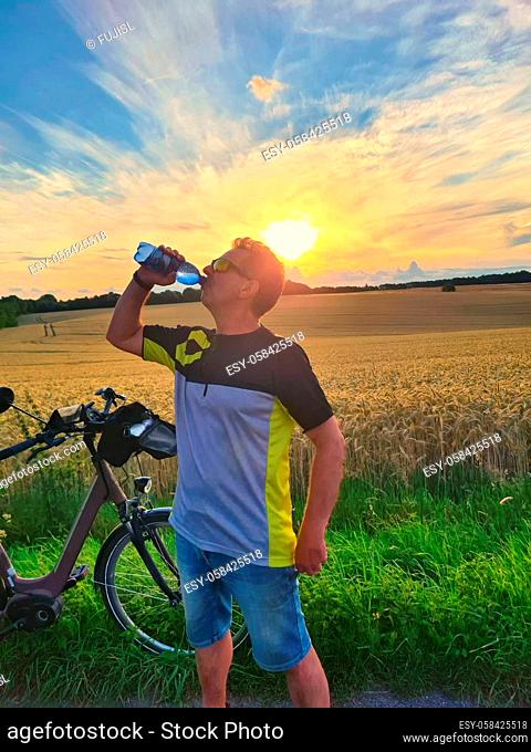 Handsome young professional cyclist dressed in cycling clothing and protective helmet feeling free and happy, drinking water from the bottle