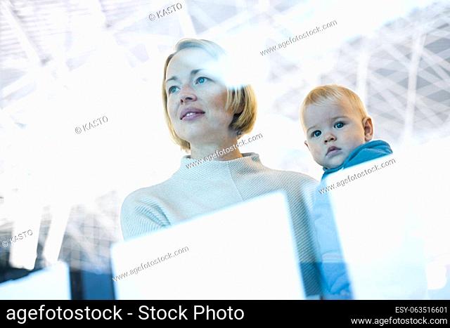 Thoughtful young mother looking trough window holding his infant baby boy child while waiting to board an airplane at airport terminal departure gates
