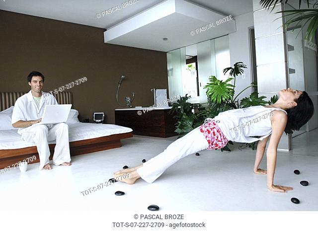 Mid adult woman practicing yoga in front of a mid adult man using a laptop