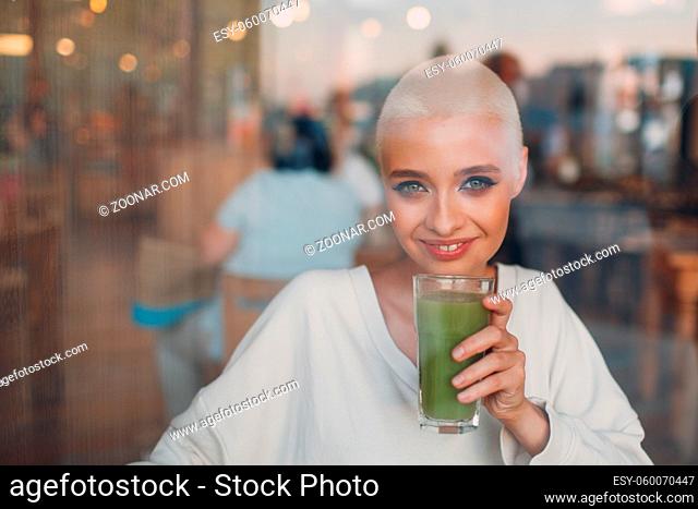 Portrait of young smiling millenial european short haired woman looking through glass with green smoothie at cafe. Beautiful happy blonde girl indoor