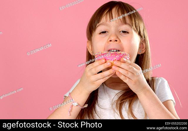 Cute young caucasian girl caught eating a sweet pink doughnut. Wearing white tshirt while standing isolated on pink background in studio