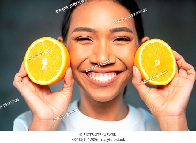 Portrait of beautiful young smiling woman in white t-shirt holding halves of orange near face over concrete background