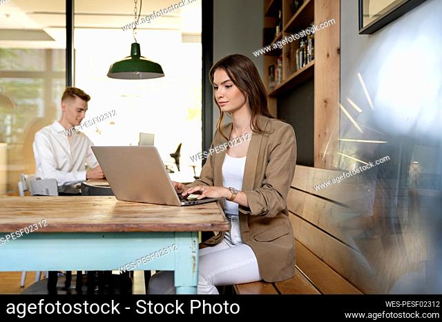 Businesswoman working on laptop while sitting with colleague in background at office