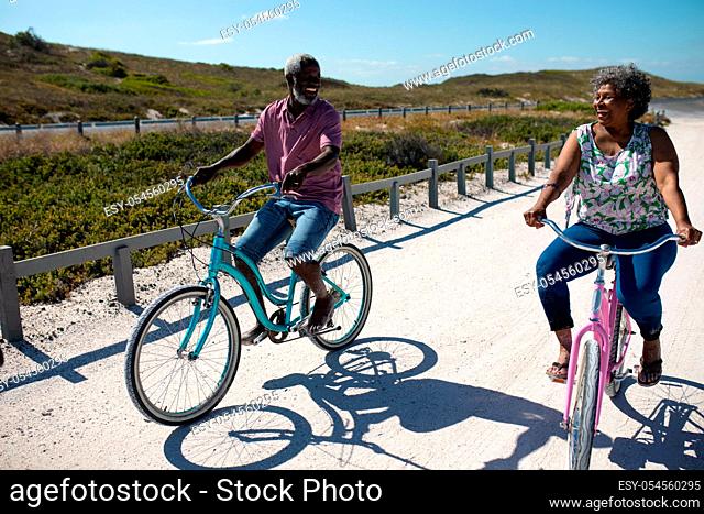 Front view of a senior African American couple riding bikes on a beach and smiling at each other, with blue sky and highlands in the background