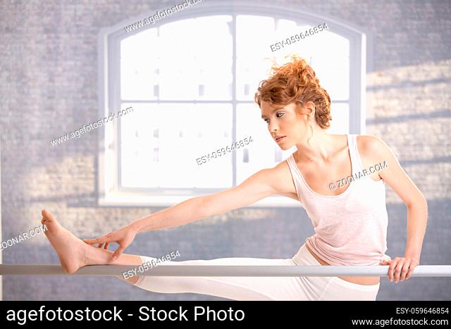 Attractive ballerina girl practicing by bar in dance studio front of window