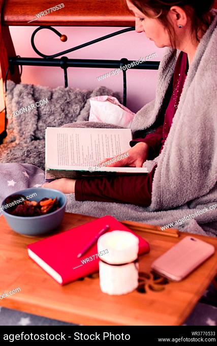 Woman enjoying the reading a book and drinking coffee at home. Young woman sitting in bed, wrapped in blanket, holding book, relaxing at home