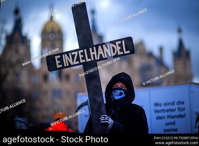 12 March 2021, Mecklenburg-Western Pomerania, Schwerin: A protester at a protest dressed in the garb of the Grim Reaper and carrying a cross with the...