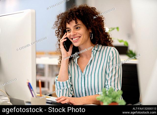 Young adult businesswoman talking on smart phone while working on computer