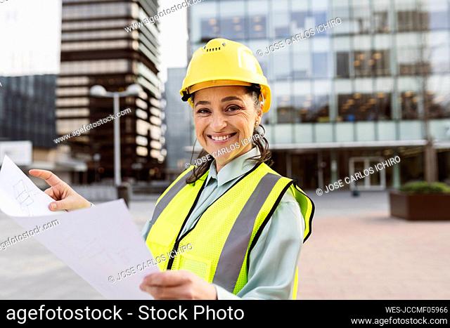 Happy female architect wearing hardhat holding blueprint