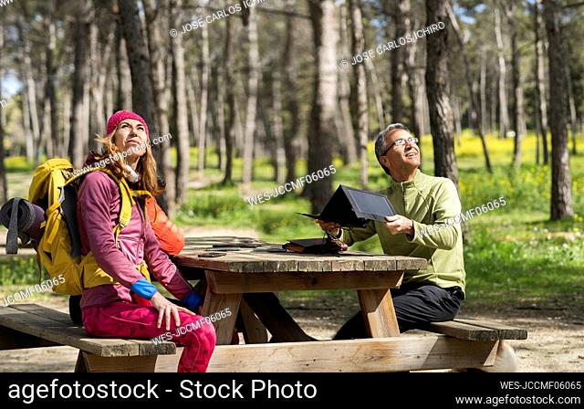 Smiling man adjusting solar panel sitting with friend at picnic table in forest
