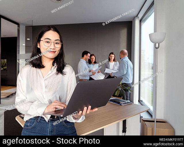 Portrait of young businesswoman using laptop with colleagues in background in office