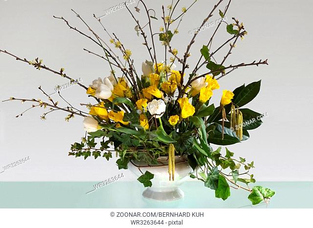 Oesterlicher Blumenstrauss mit gelben Osterglocken, weissen Tulpen, Zweigen und Efeuranken in einer weissen Terrine auf einem Glastisch. Querformat