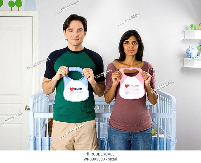 Portrait of a couple standing in front of a crib and holding baby bibs