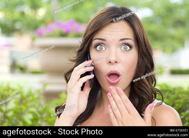 Shocked young adult female talking on cell phone outdoors on bench
