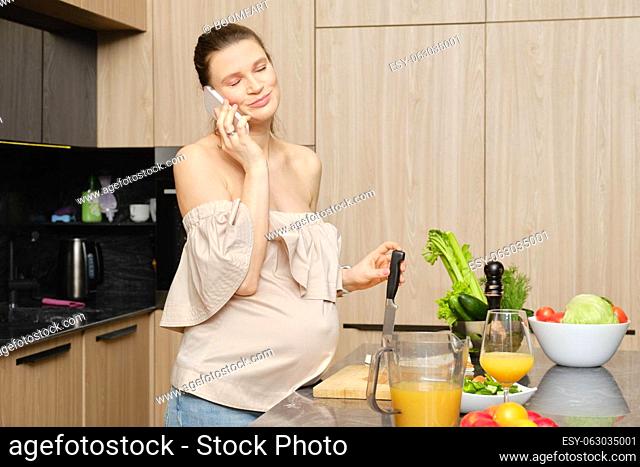 Pregnant woman woman leans on a kitchen knife and talks on the phone. Woman closed her eyes listening pleasant news during phone conversation