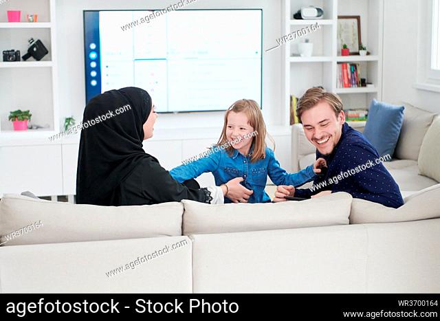 Happy Muslim family with daughter woman in traditional fashionable dress having fun and good time together while sitting on sofa during the month of Ramadan at...
