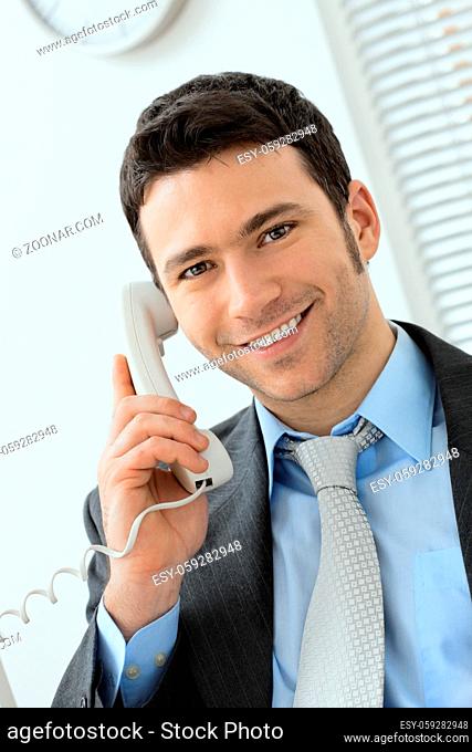 Happy young businessman talking on landline phone at office, smiling
