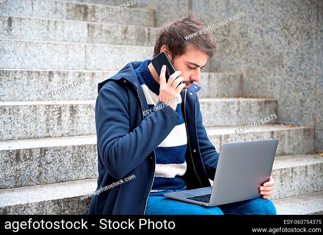 Man talking on phone while working on laptop sitting in stair steps in urban scenery
