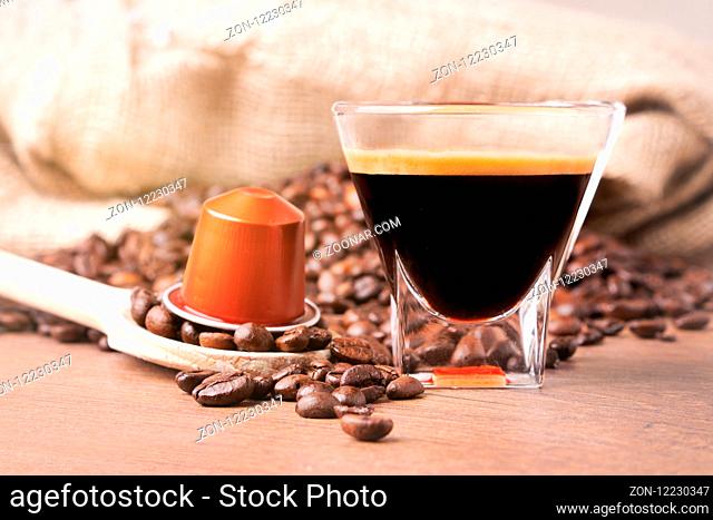 Cup of coffee with coffee capsule on wooden spoon, roasted coffee beans on wooden background, front view