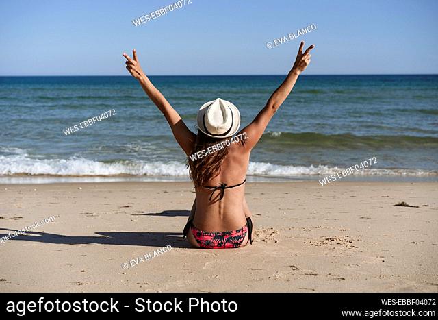 Carefree woman gesturing peace sign while sitting on beach