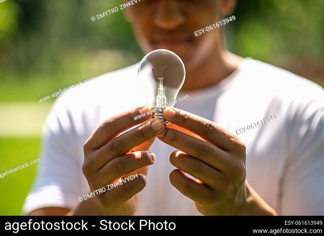 Reflections. Hands of dark-skinned guy standing in nature on sunny dayshowing light bulb at camera